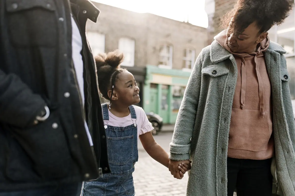 a young girl in overalls holding hands with her parents on a sidewalk