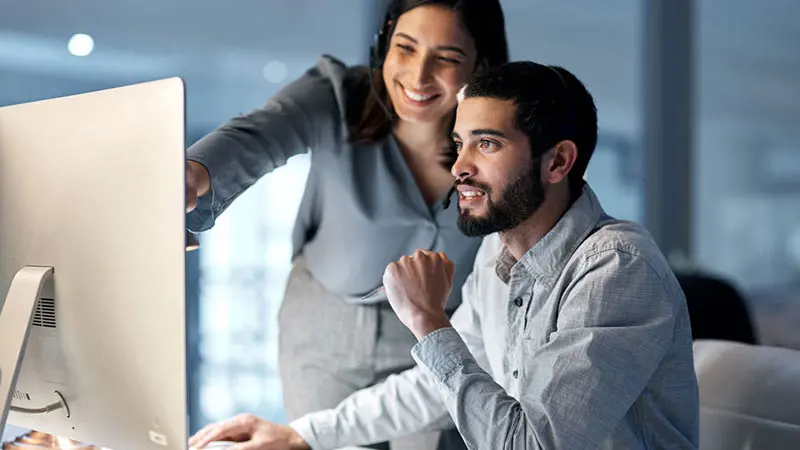 Two young professionals, one male and one female, who are looking at a computer monitor together as they discuss the order to cash cycle