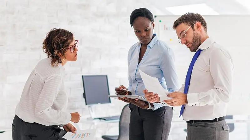 Three professional colleagues standing in an office discussing the monthly economic situation