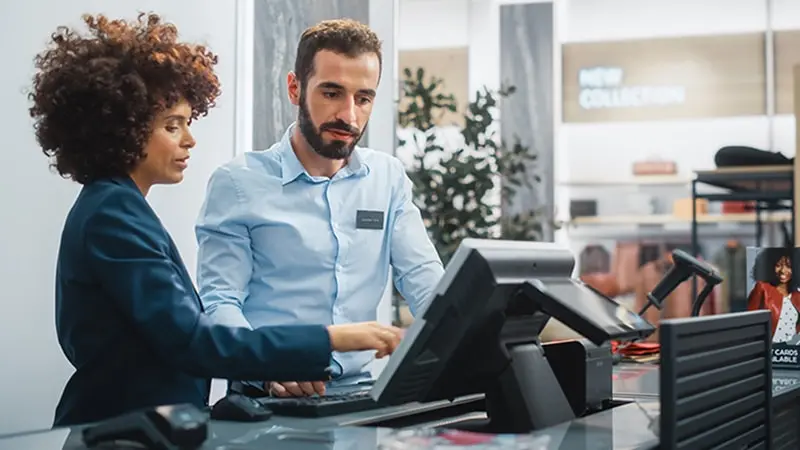 One retail worker in a department store showing another worker how to use the point of sale machine