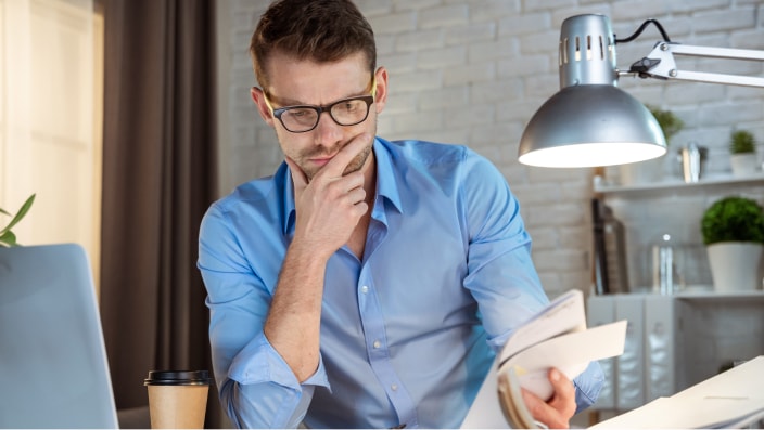 Man wearing glasses and a blue button down shirt, sitting at a desk leafing through paperwork.