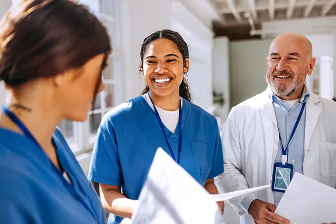 Three U.S. healthcare workers in blue scrubs and lab coats smiling while discussing the healthcare economy.