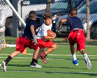 Children playing flag football on a turf field, with one child carrying a football while teammates move to defend.