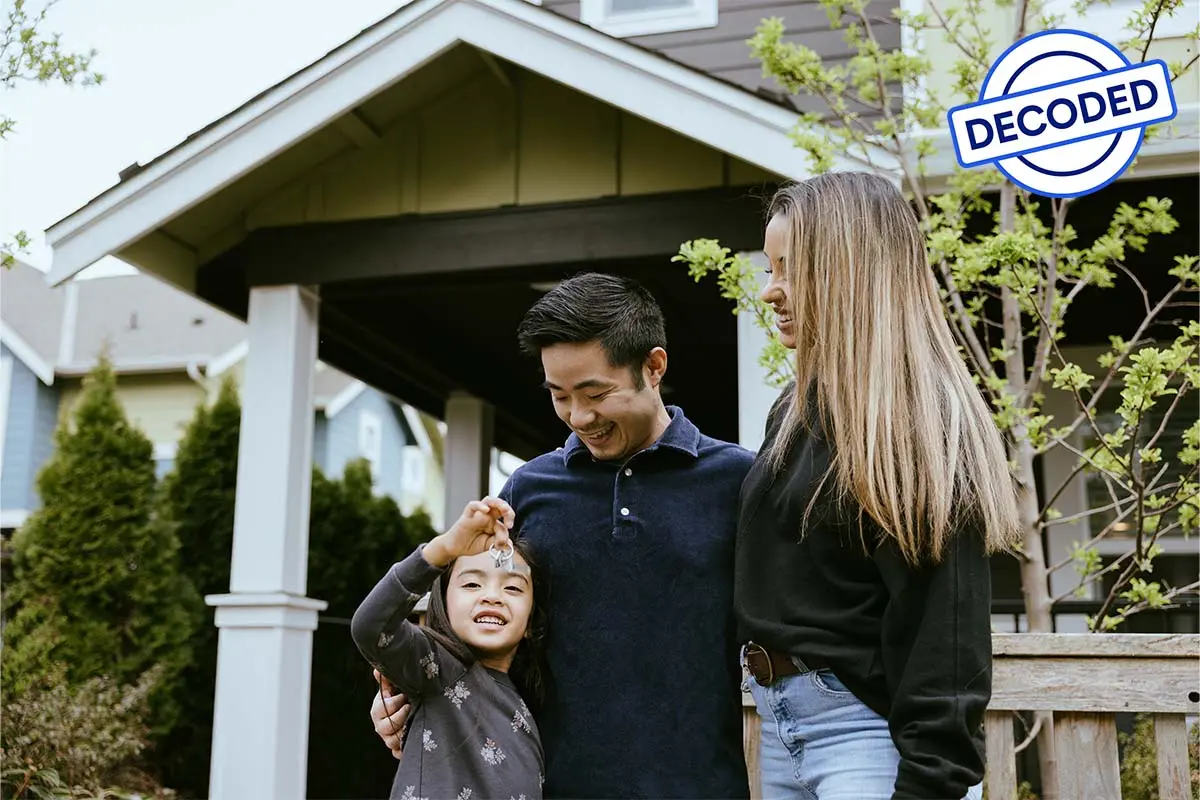 A family of three stands outside a house, with a girl holding keys, smiling