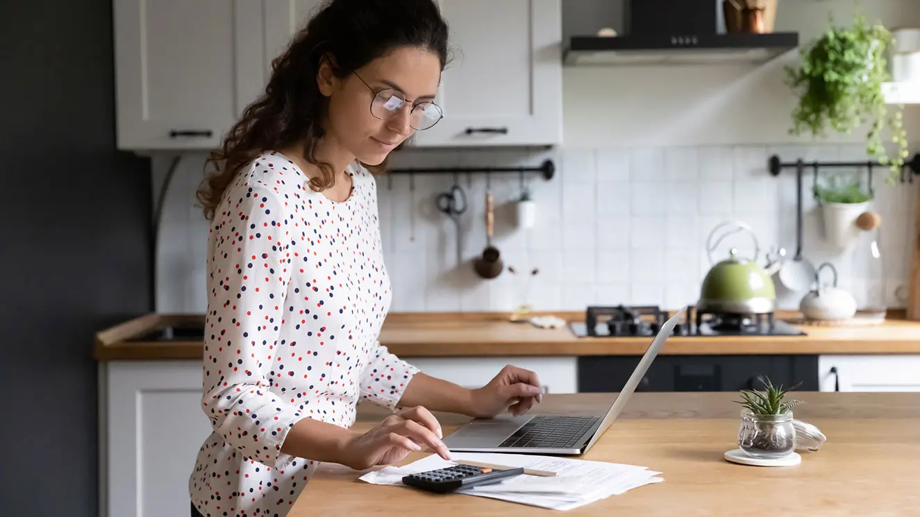 woman standing at her kitchen island managing her money on her computer