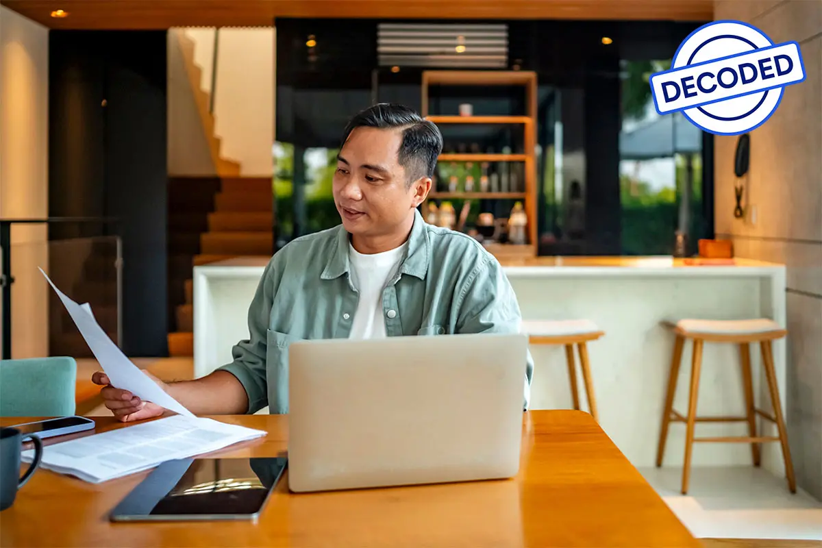 A man in a green shirt sits at a wooden table with a laptop and paperwork, looking focused.