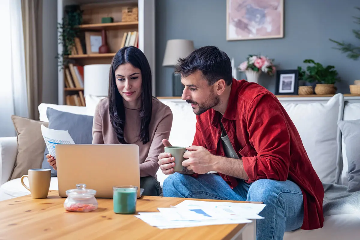 A man and a woman sitting on a couch, looking at a laptop with printed papers and drinks on the table.