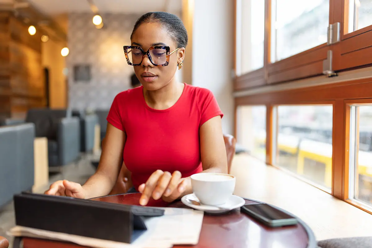 A woman in a red shirt wears large sunglasses, using a tablet with coffee nearby