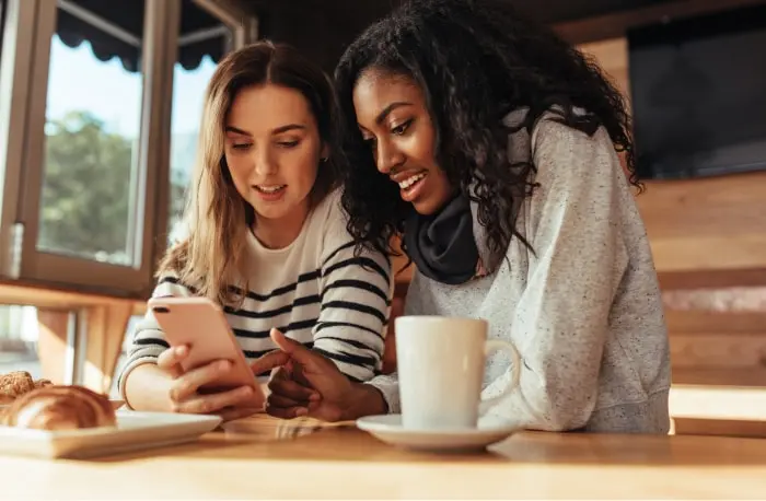 Two friends at a restaurant table leaning in toward a phone with one holding it while the other is pointing at the screen