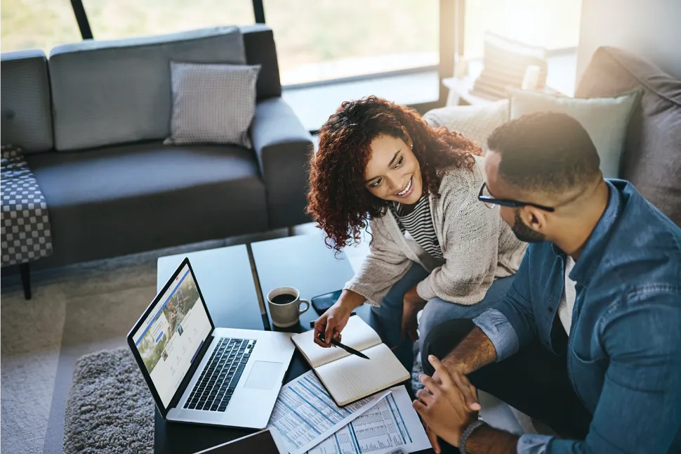 A young couple sitting on their sofa and looking at a laptop while writing down notes about their retirement plan  