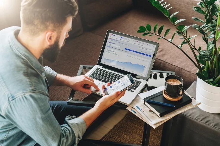 Bearded younger man looking at stock charts on his laptop and phone