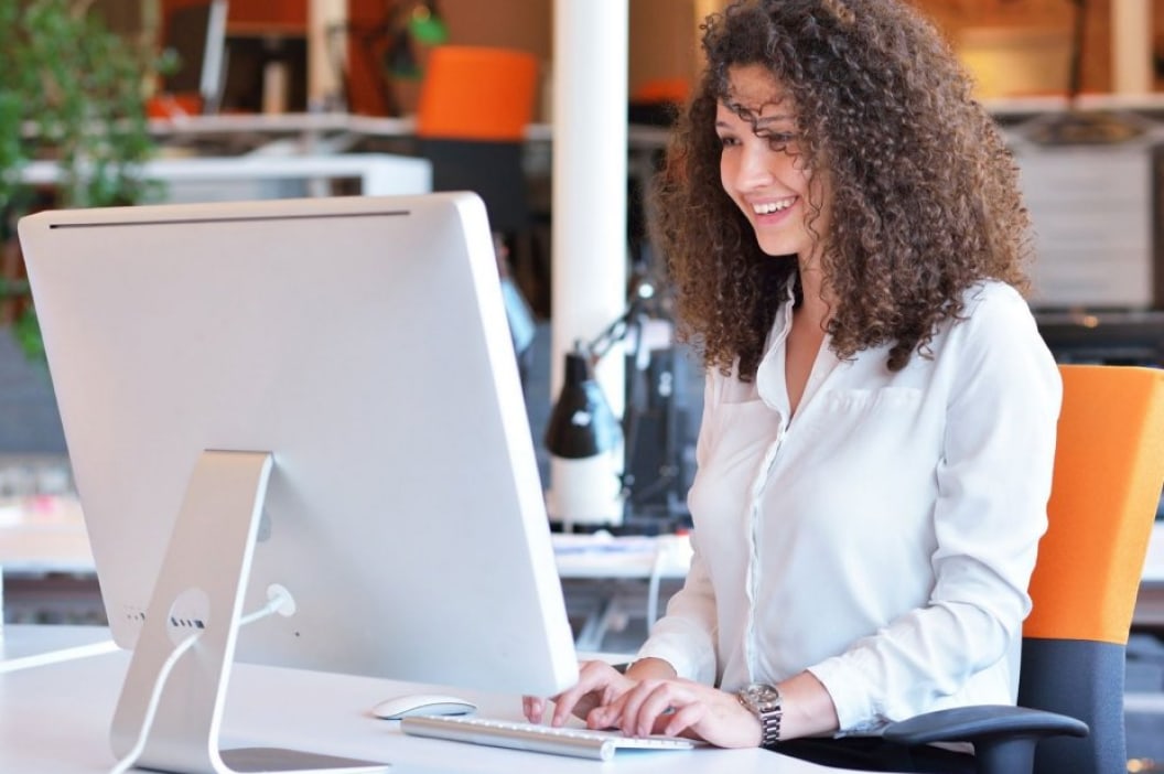 Young woman in an open, modern office sitting in front of a large computer monitor to do work.